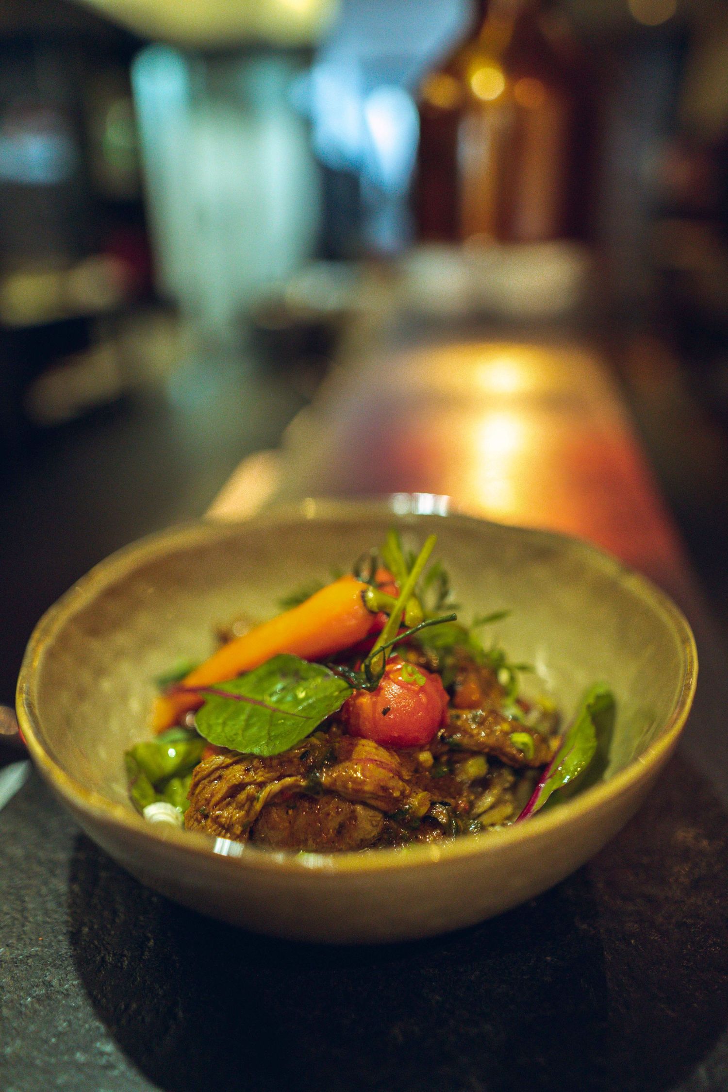 Gourmet dish served in earthen bowl with roasted meat, fresh vegetables, and herbs in warm ambient lighting.