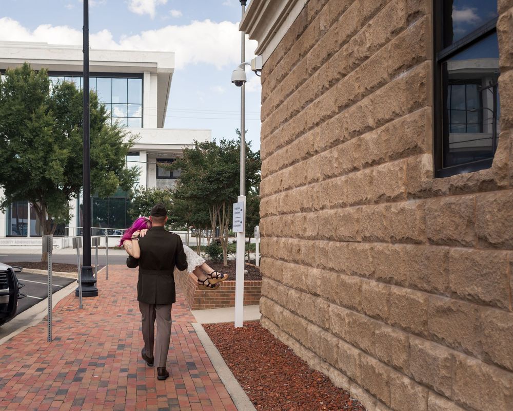 Person in suit carrying pink item walks along brick sidewalk beside historic stone building in urban setting.
