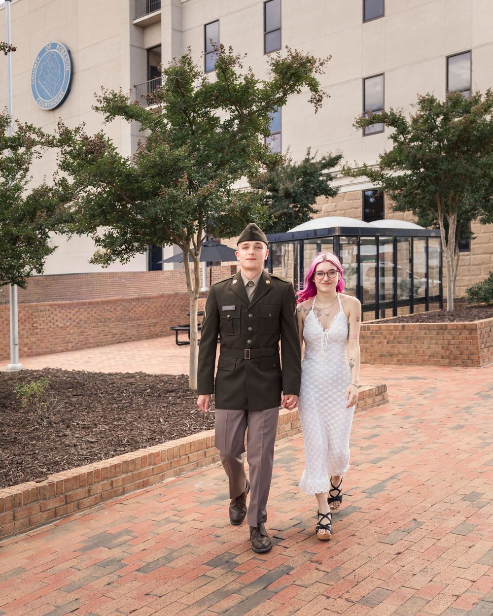 Military man in uniform walks hand-in-hand with pink-haired woman in white dress on brick plaza.