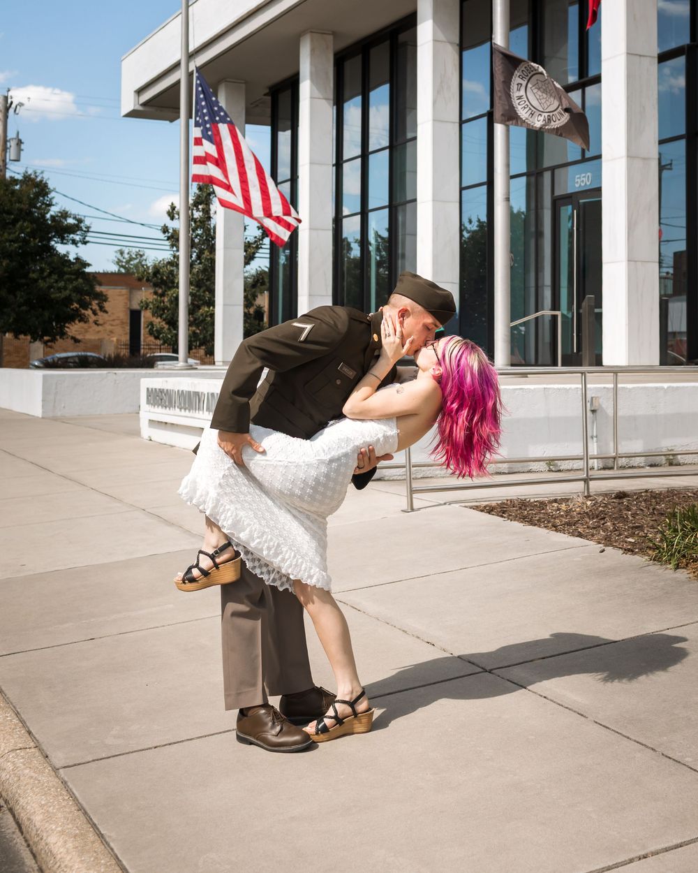 Couple recreating iconic WWII kiss pose in front of American flag, woman with pink hair in white dress.