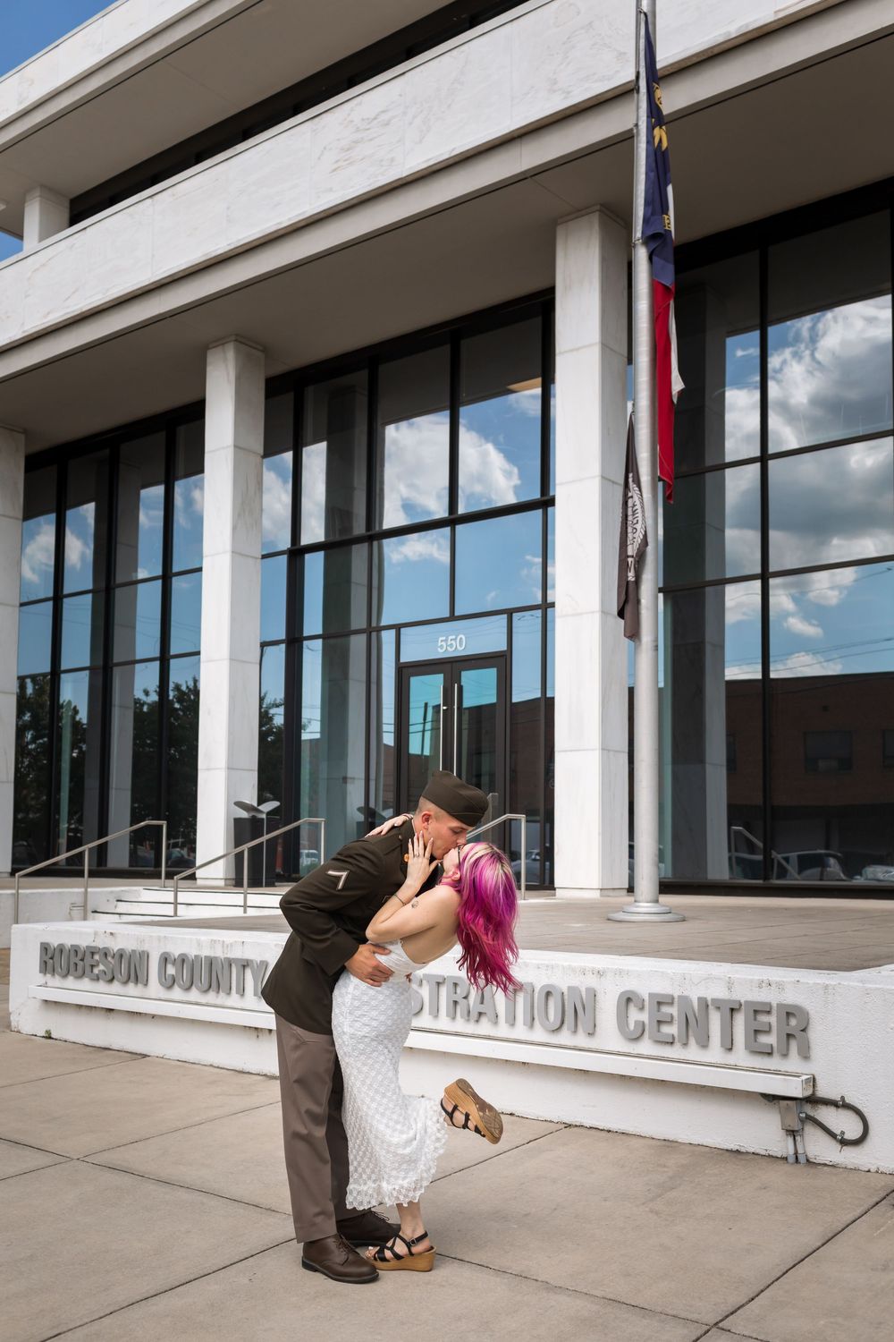 Couple sharing a kiss in wedding attire outside Robeson County Administration Center with flags in background.