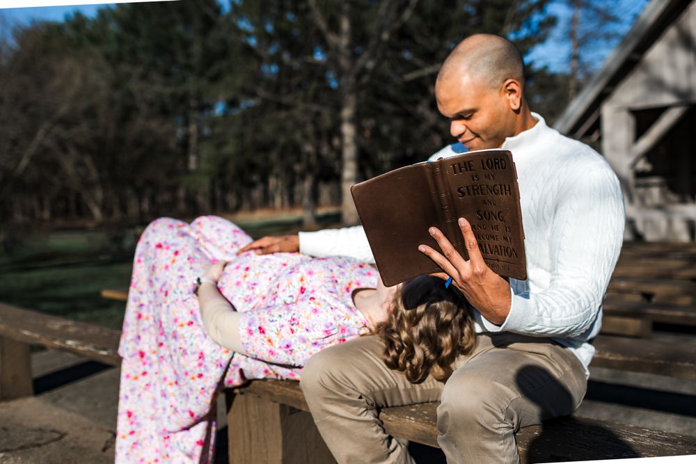 Man in white turtleneck reads a book to a child outdoors on a sunny day near a rustic wooden structure.