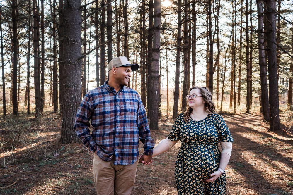 Couple holding hands in a sunlit forest during a maternity photo session, woman in floral dress touching baby bump.