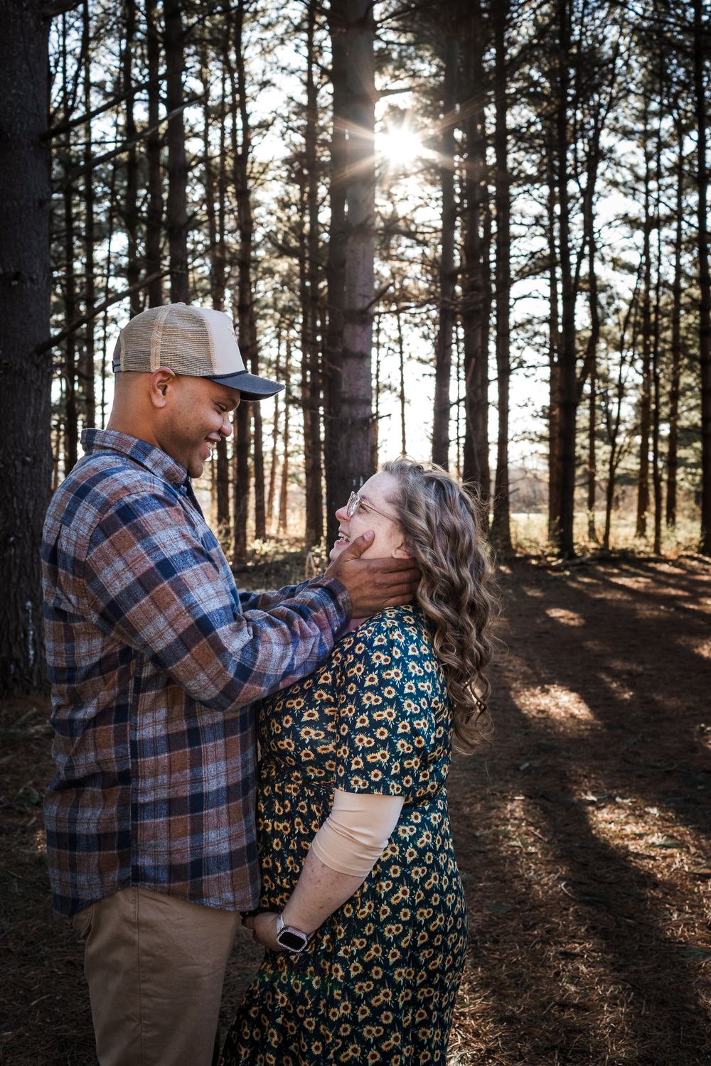 Couple sharing a romantic moment in a sunlit pine forest, man in plaid shirt and cap gently holding woman's face.
