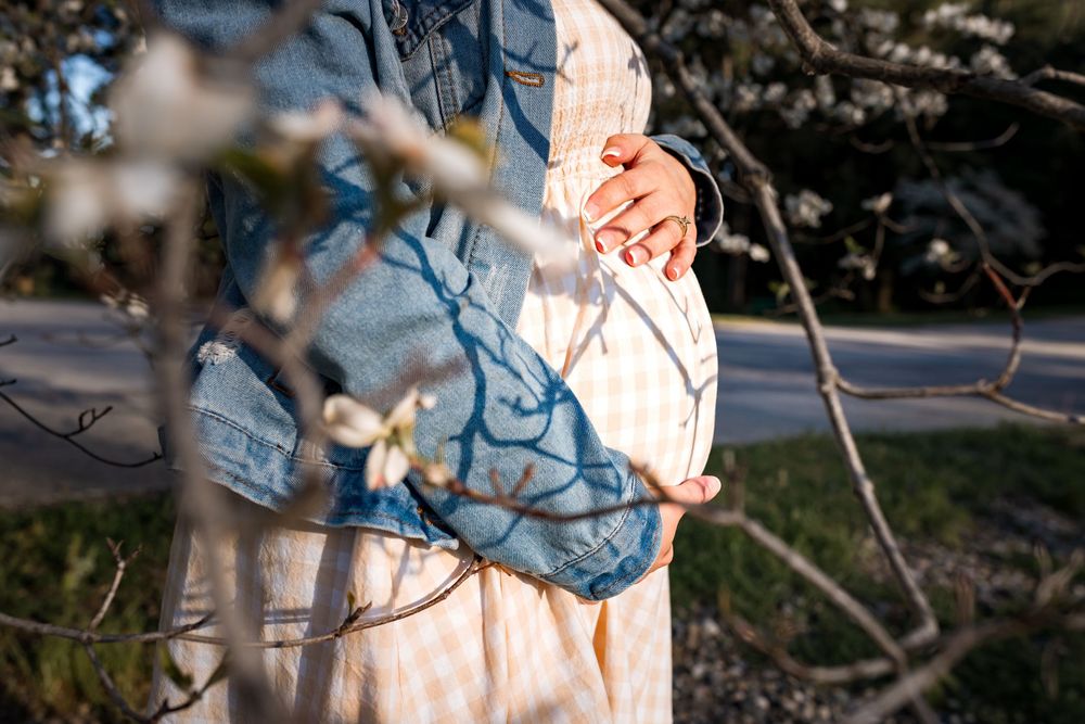 Pregnant woman in denim jacket and floral dress cradling baby bump outdoors near blossoming tree branches.