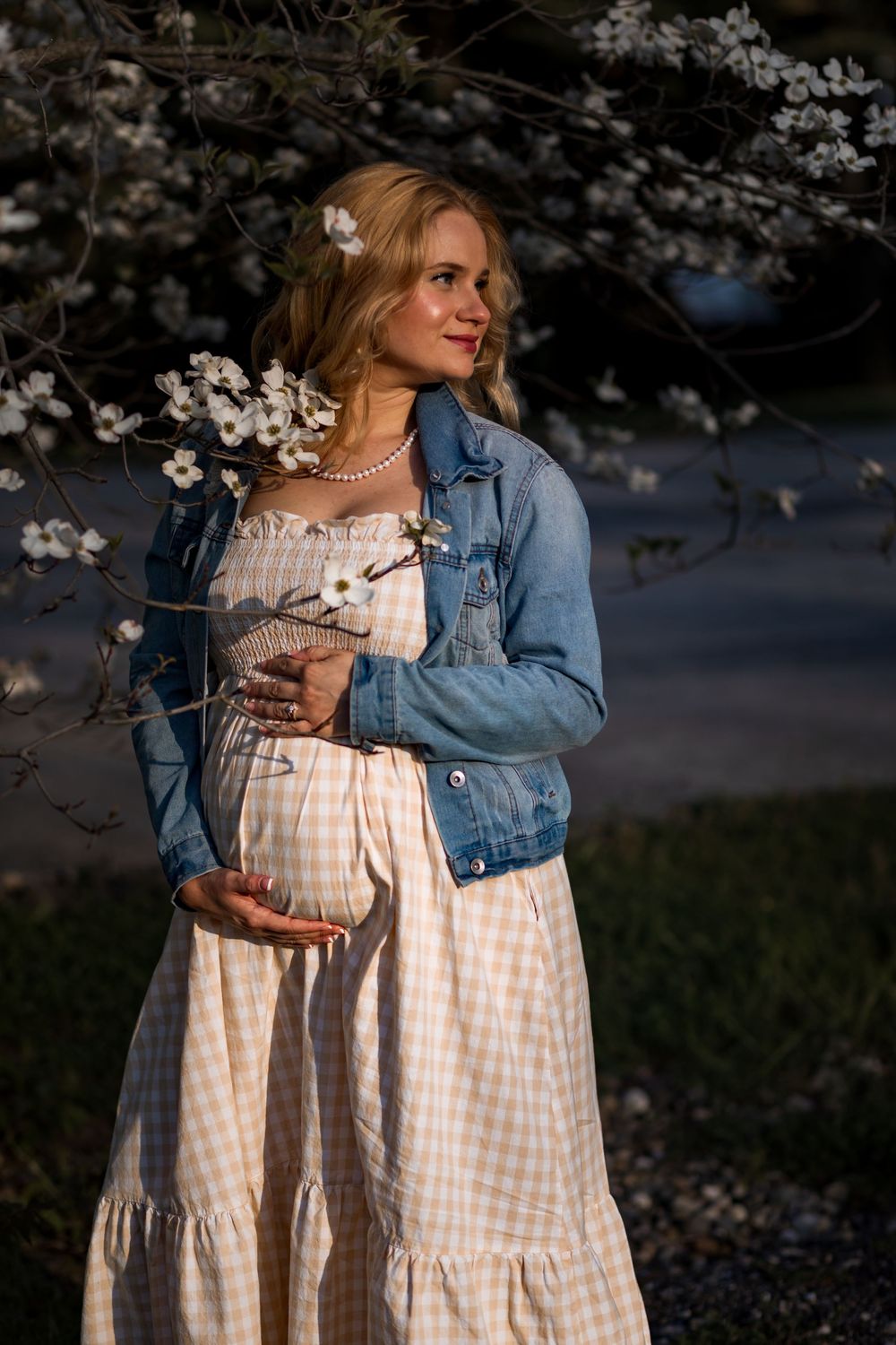 Pregnant woman in a plaid dress and denim jacket standing beside a flowering dogwood tree at sunset.