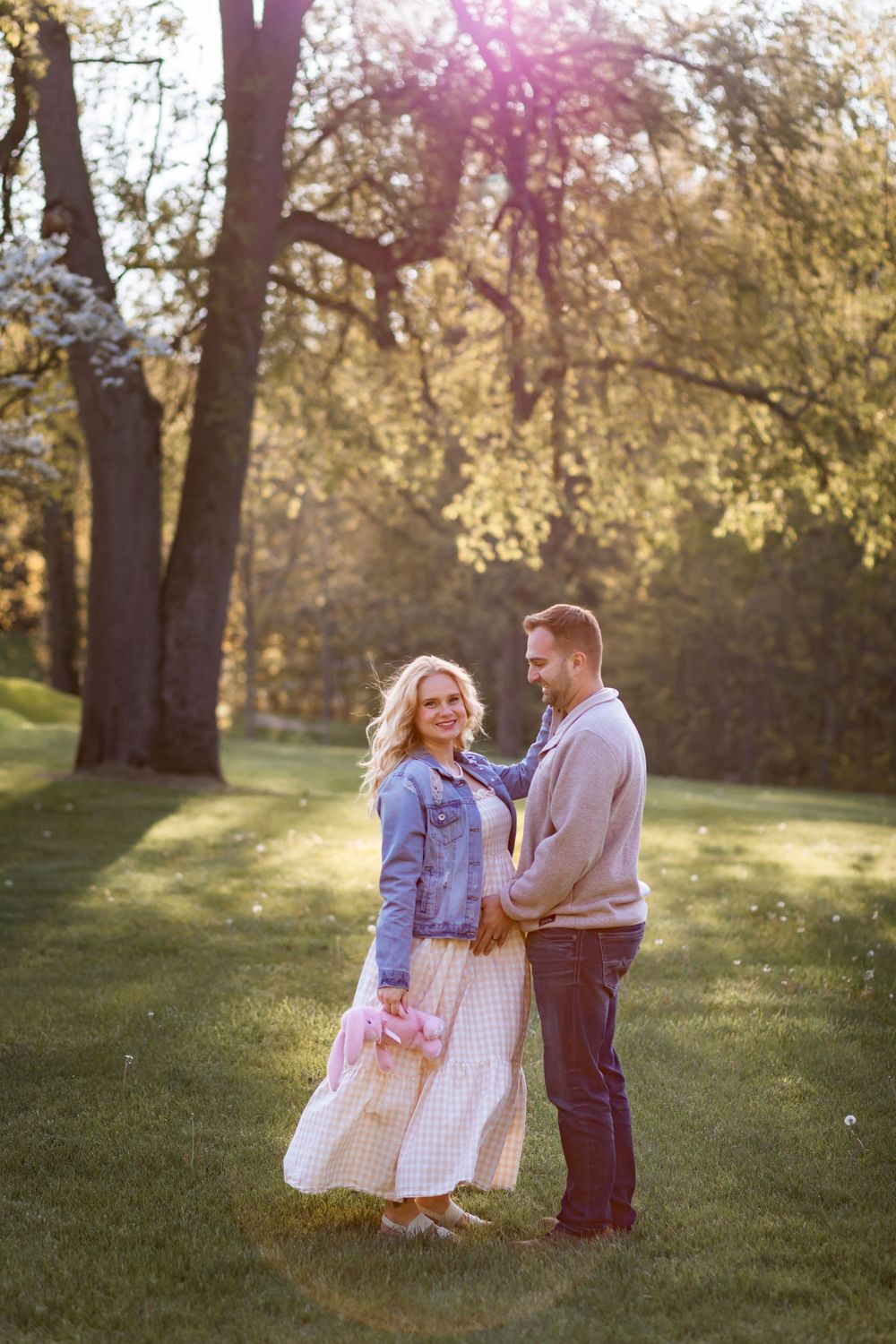 Couple embracing in sunlit park during golden hour, woman in denim jacket holding pink flowers, surrounded by blooming trees.