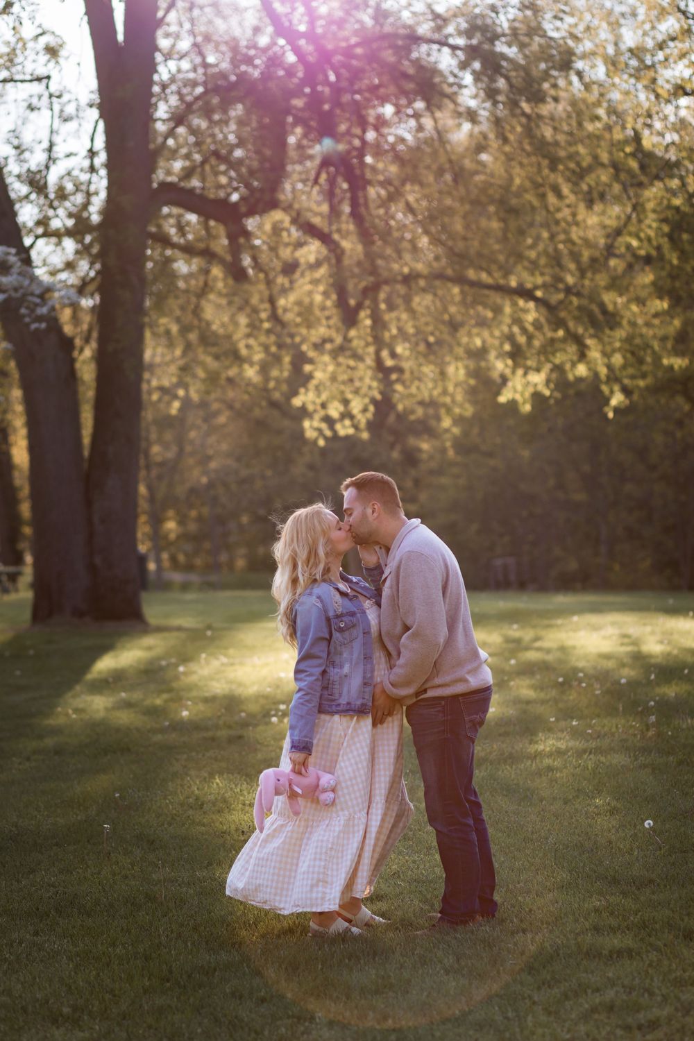 Couple kissing in a sunny park, woman in denim jacket and floral skirt, man in beige sweater, golden hour light.