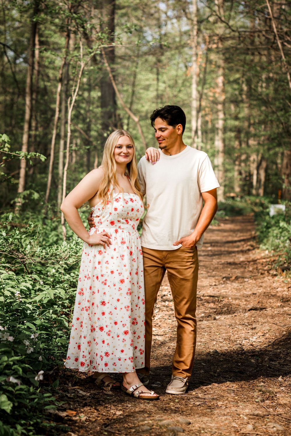 Couple posing on a forest trail, woman in floral dress and man in white tee with tan pants.