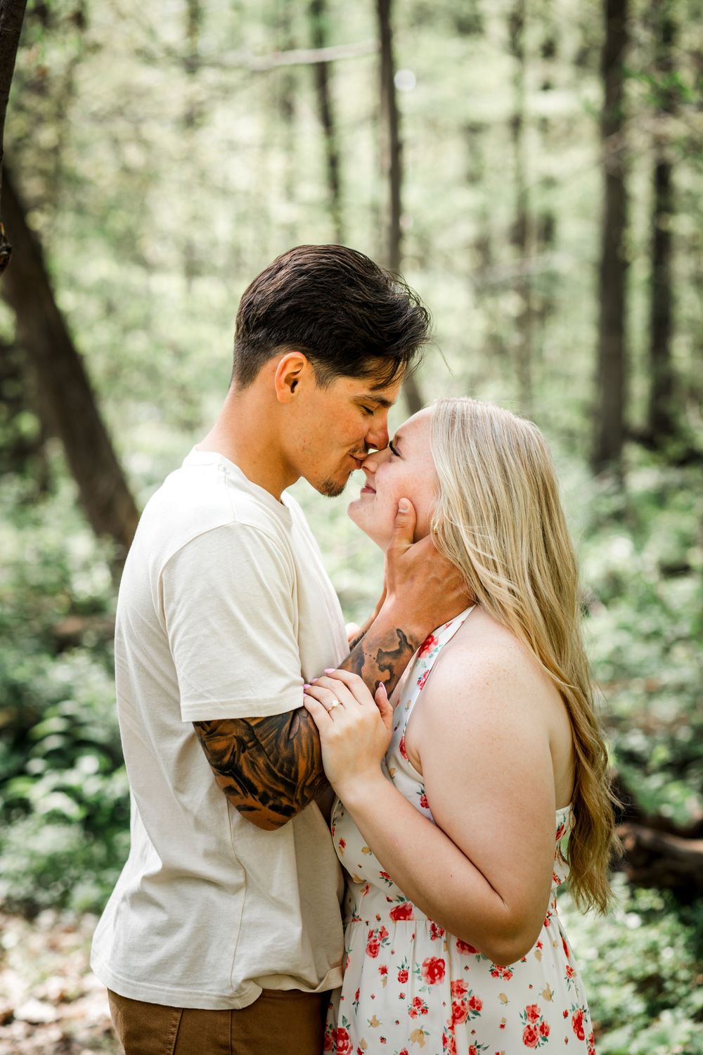 Romantic couple almost kissing in a lush green forest setting, man with tattoos in white tee.