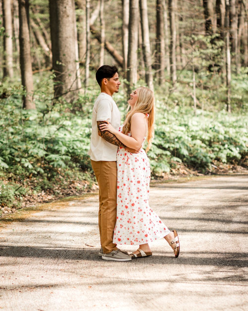 Couple embracing on a forest path, woman in floral dress and man in tan pants, surrounded by lush green trees.