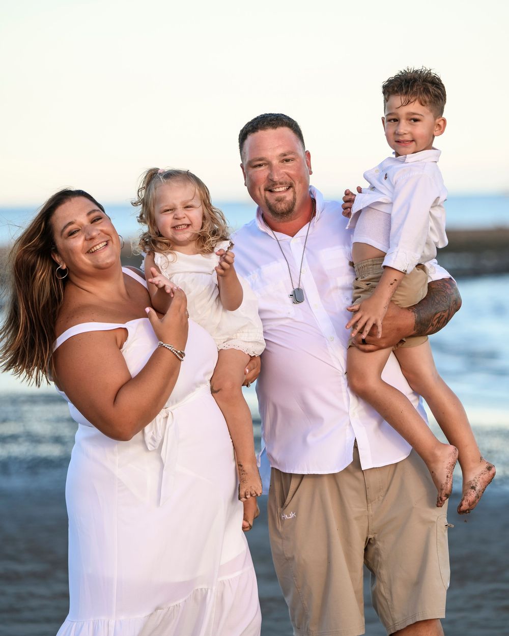 Happy family of four in white clothing posing on a beach at sunset, parents each holding a child.