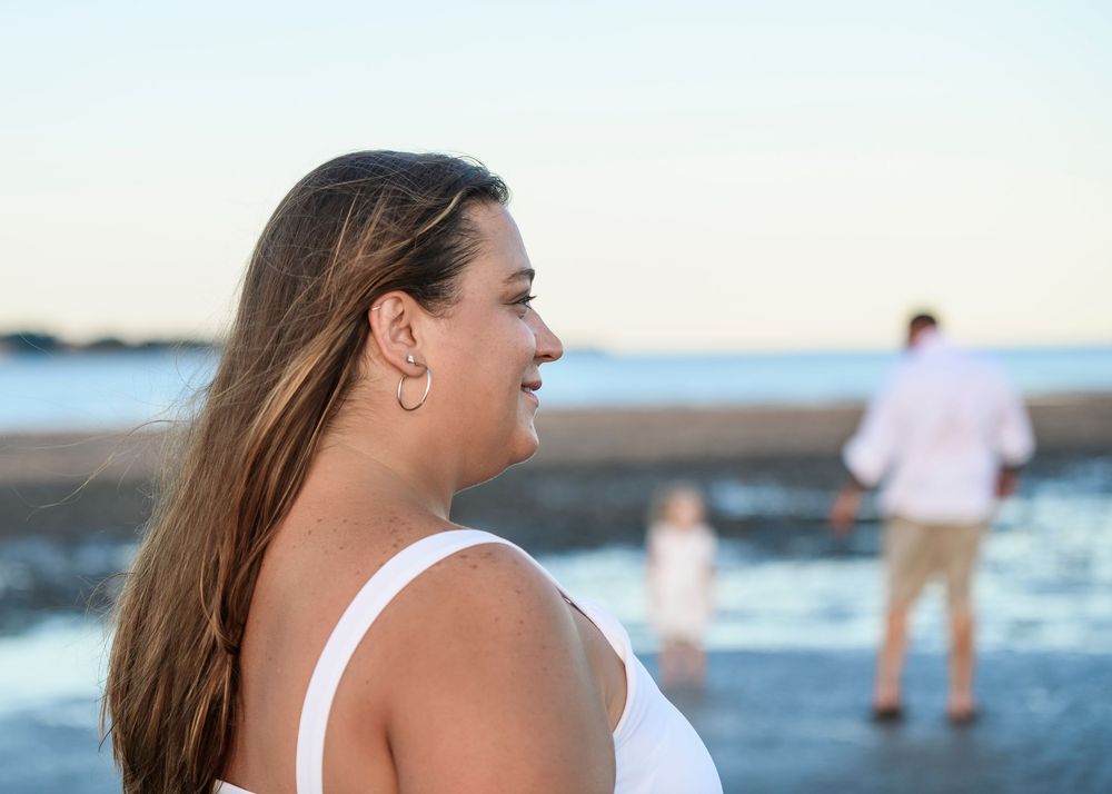Woman in white dress smiling on a beach with family in the background near the ocean shoreline.
