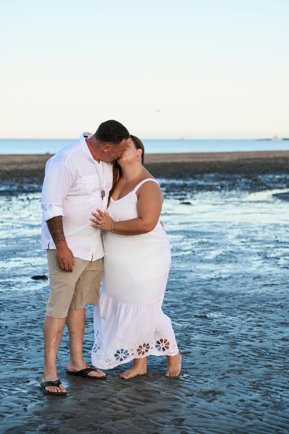 Couple sharing a romantic kiss on a wet beach at sunset, both dressed in white clothing.