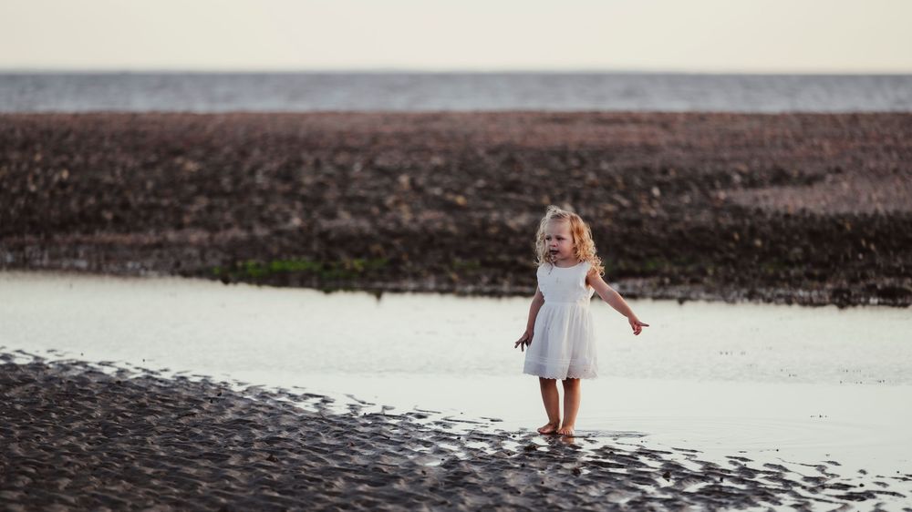 Young girl in white dress walking alone on a sandy beach near the water's edge at low tide.