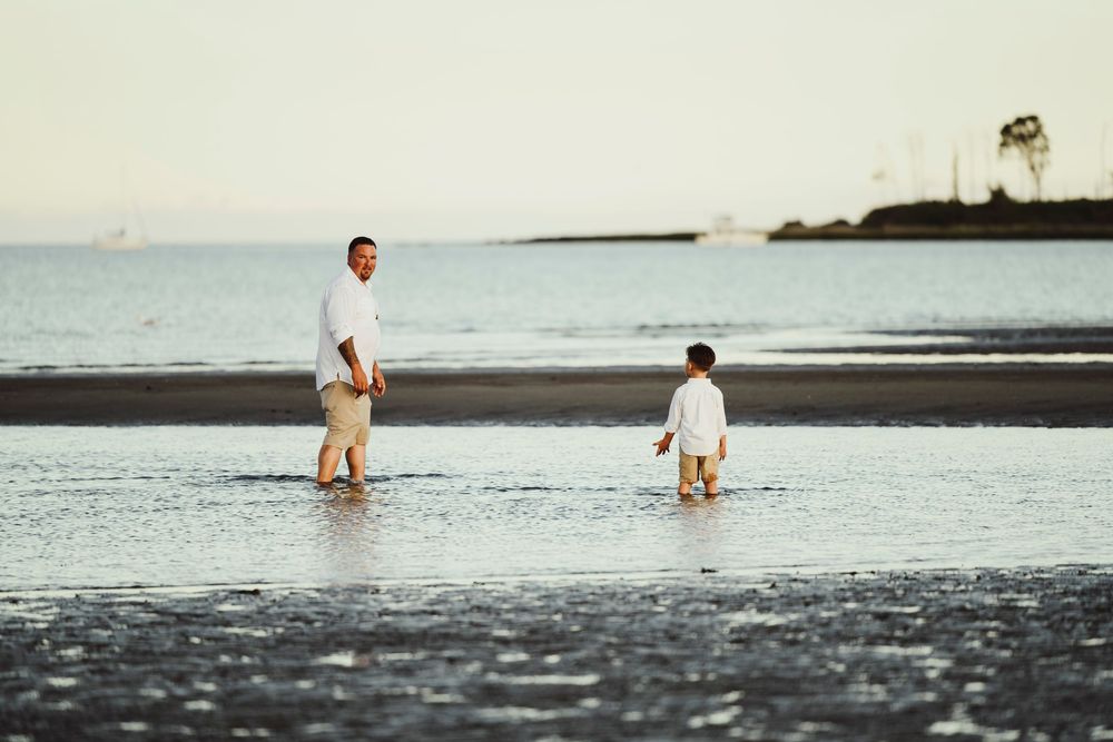 Father and young son wading together in shallow ocean water on a calm beach at sunset.
