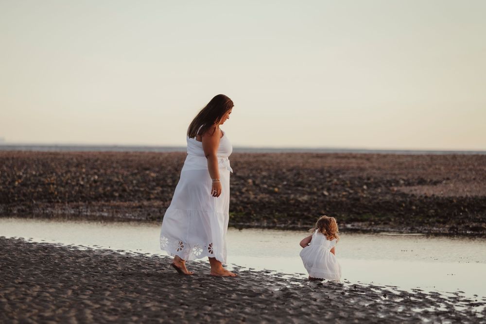 Mother and young daughter in matching white dresses walking together along a serene beach shoreline.