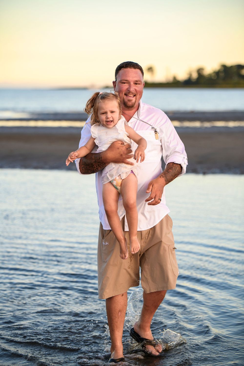 Smiling father in white shirt holding his laughing young daughter while standing in shallow beach water.