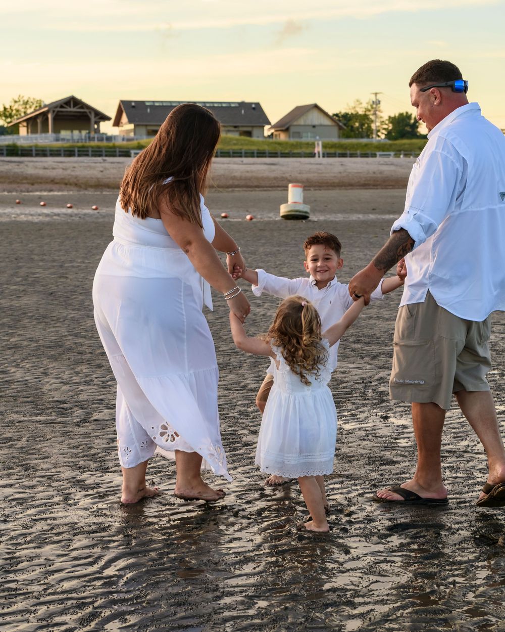 Family of four in white outfits holding hands and walking together along a wet sandy beach.