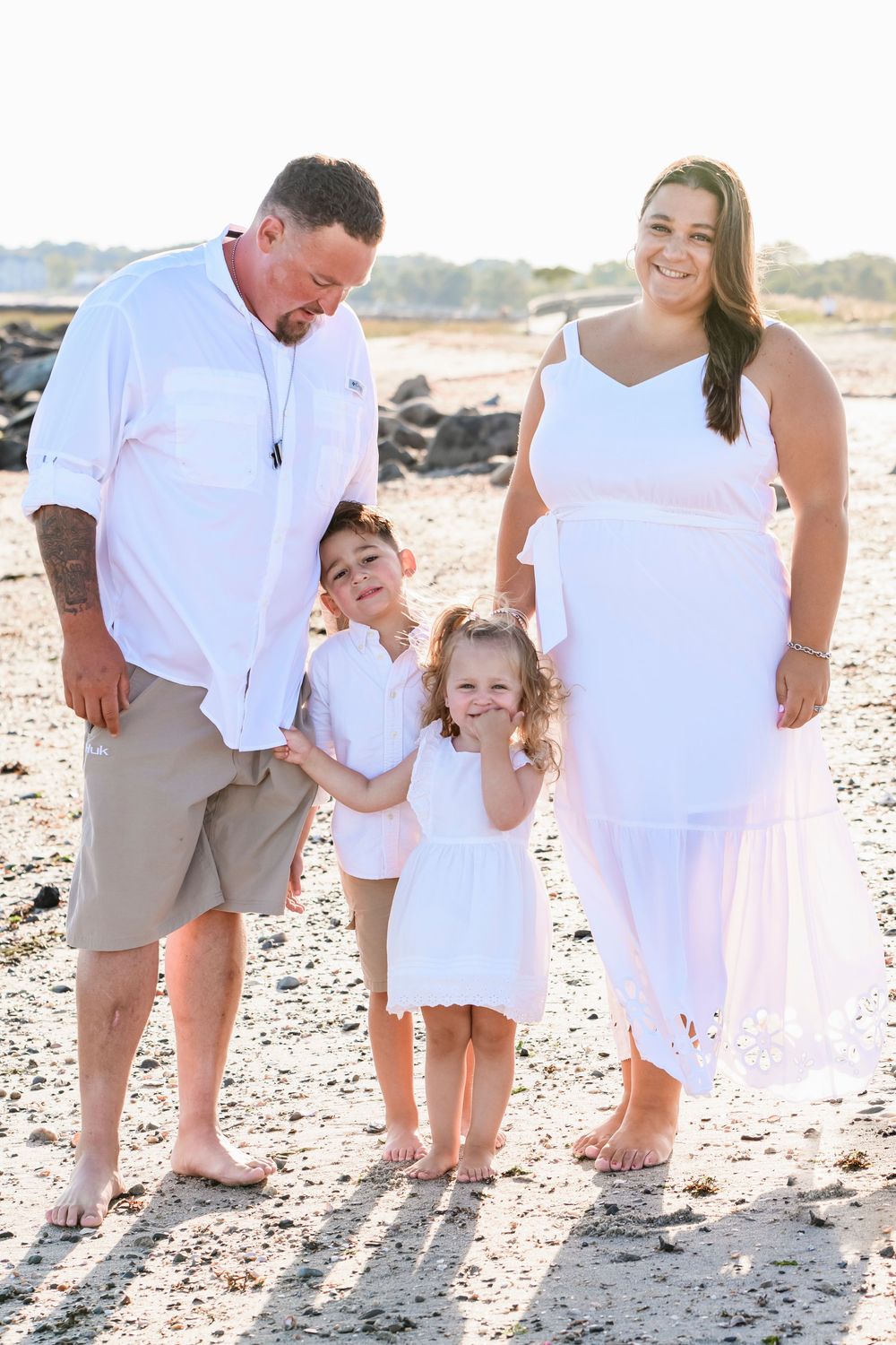 Family of four in white outfits posing on a sunny beach, parents smiling at their two young children.