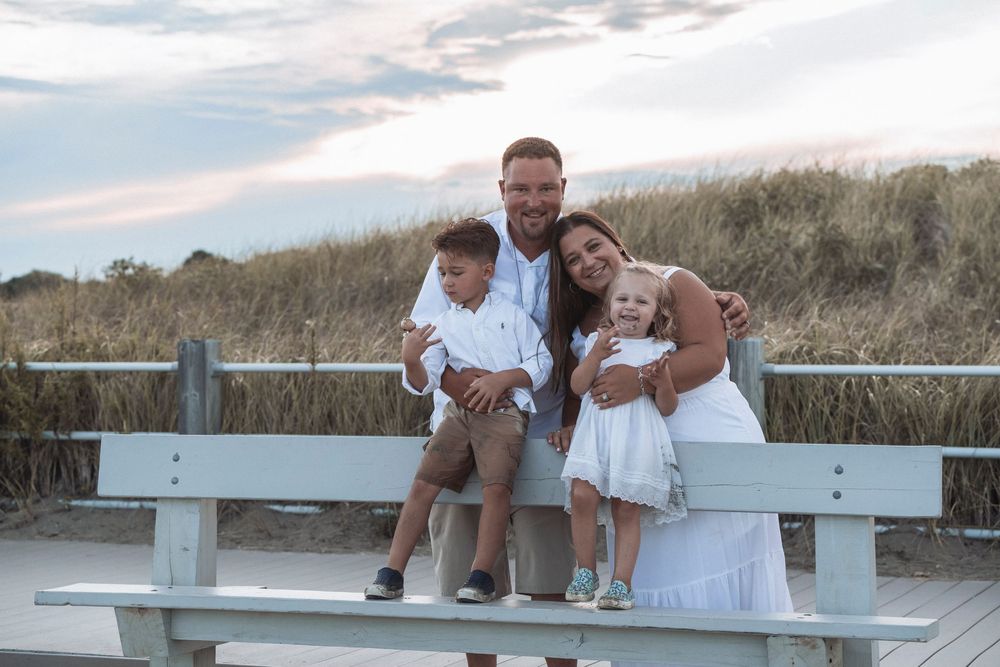 Family of four in white outfits sitting together on a light blue bench near beach dunes.