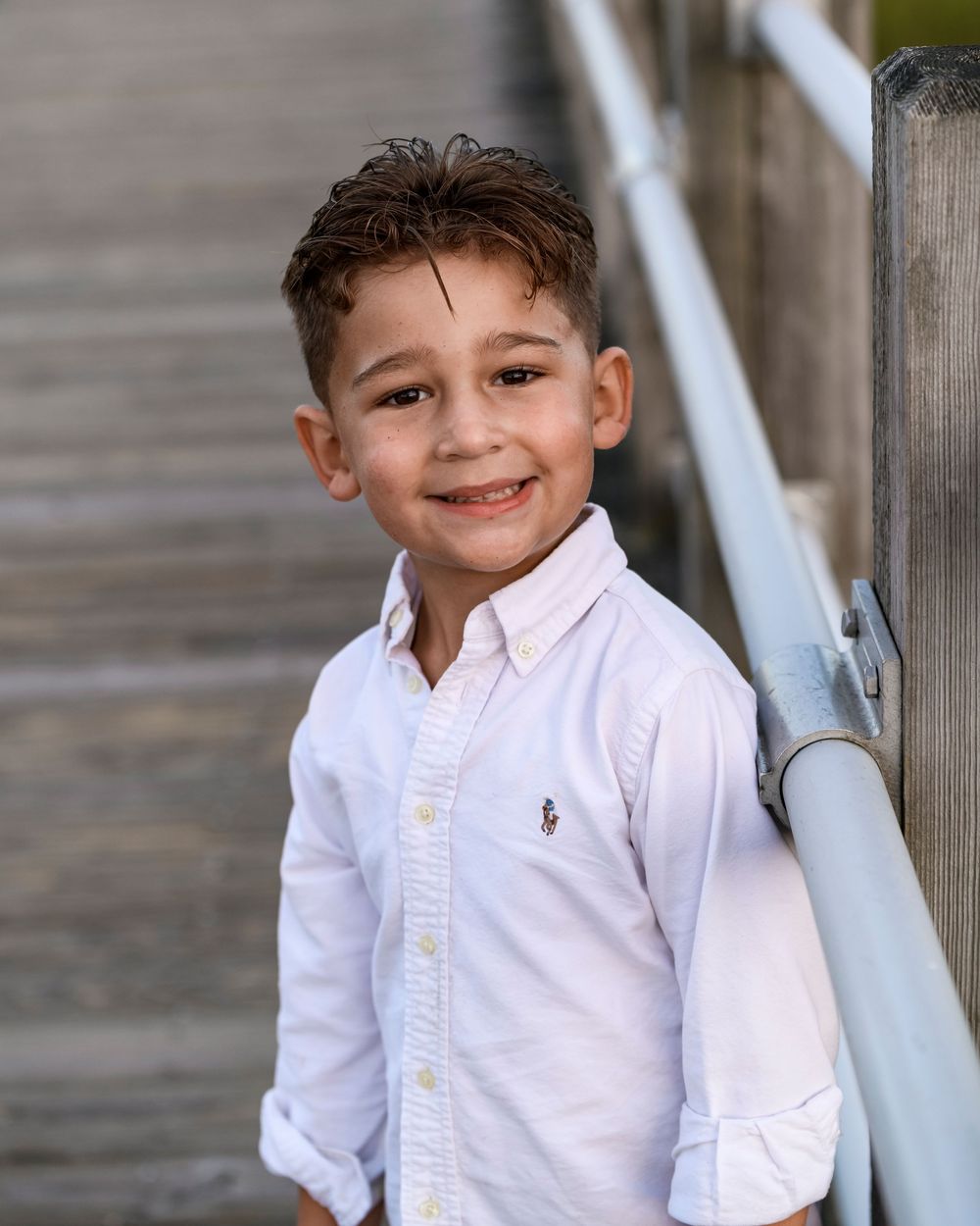 Smiling young boy in white button-down shirt leaning against railing outdoors.