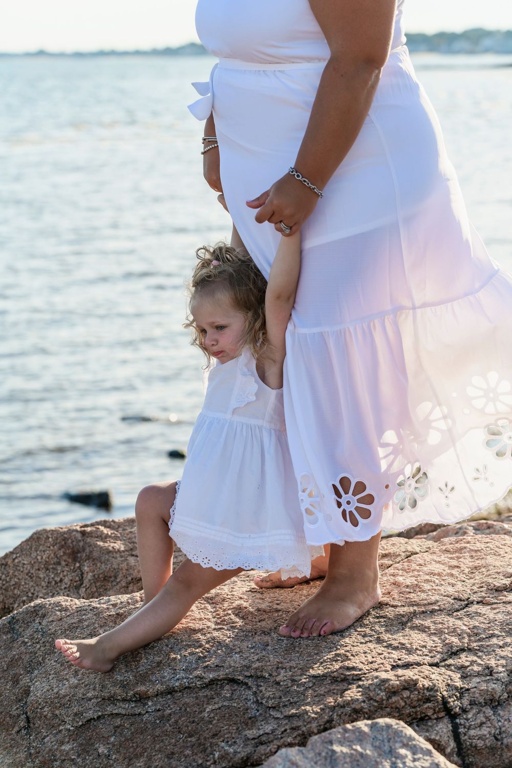 Little girl in white dress holds mothers hand while standing on rocks by the sea.