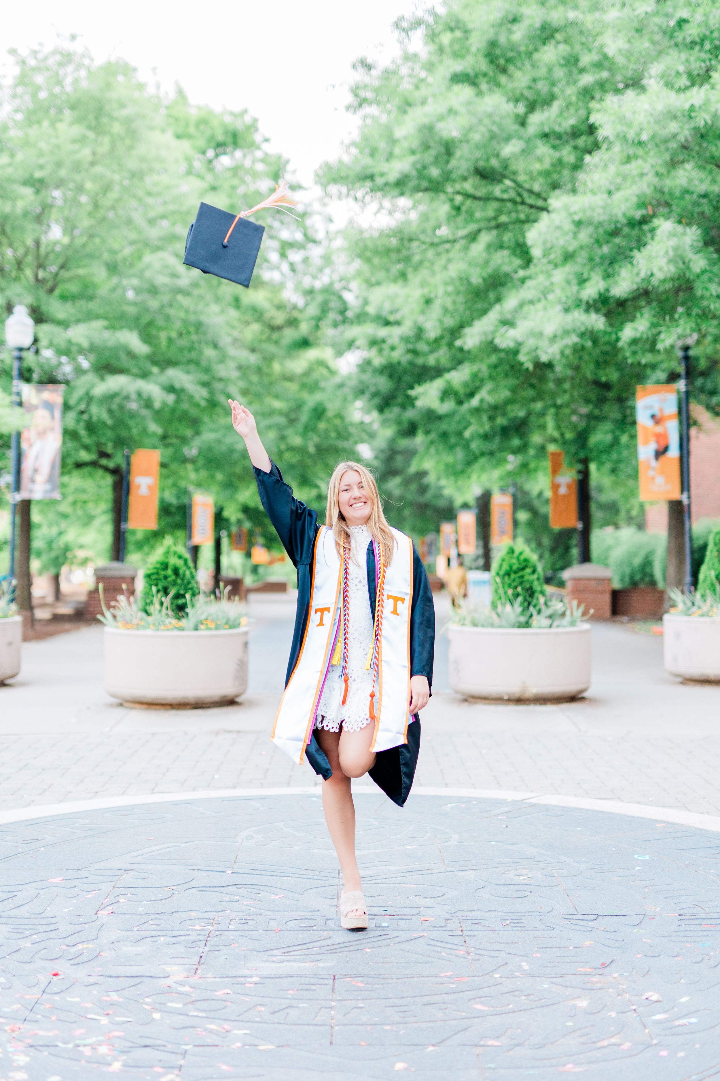 Graduate celebrates on brick walkway surrounded by green trees and orange banners during outdoor graduation ceremony.