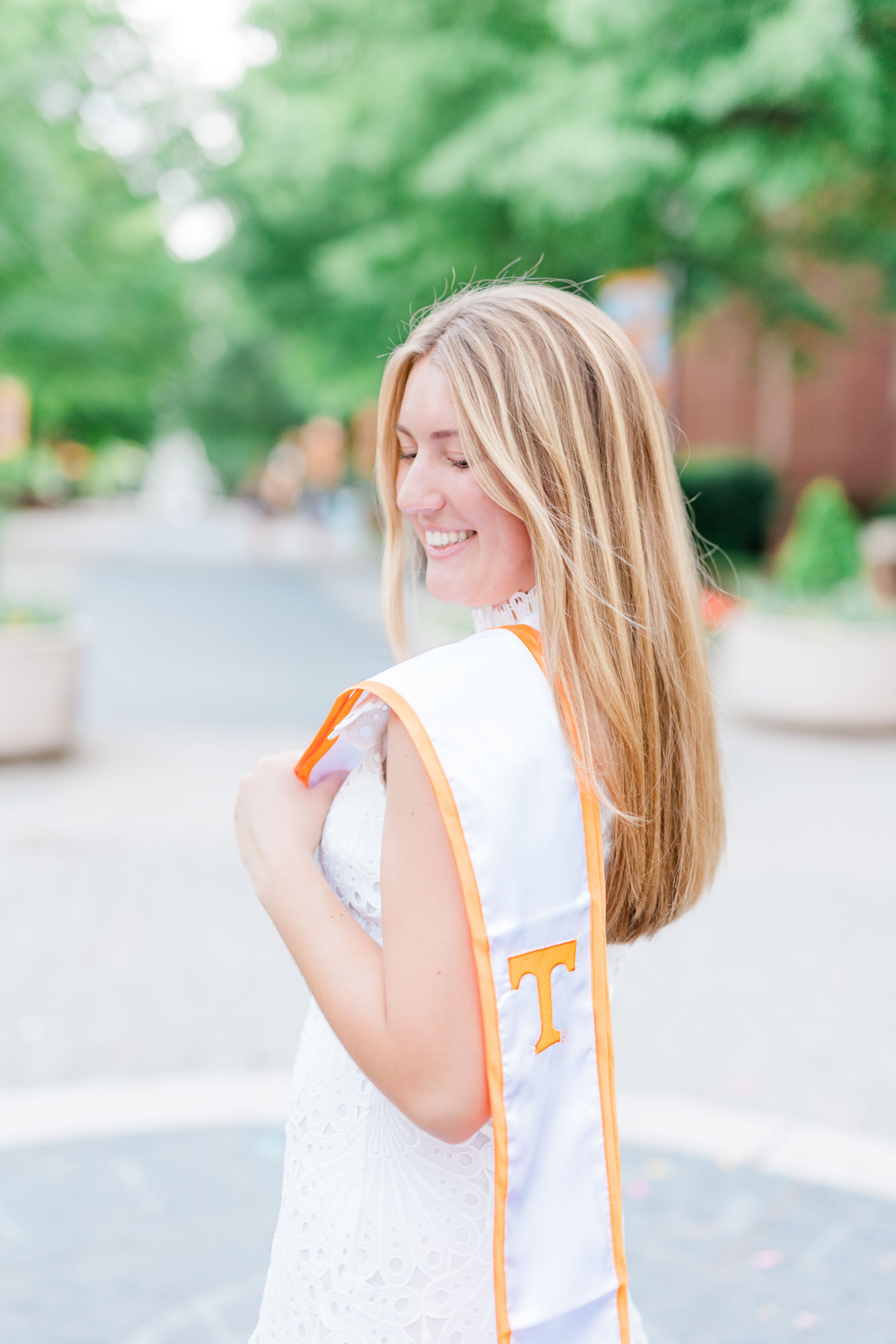 A graduate wearing a white dress and orange graduation stole with a T emblem poses outdoors on a sunny day.