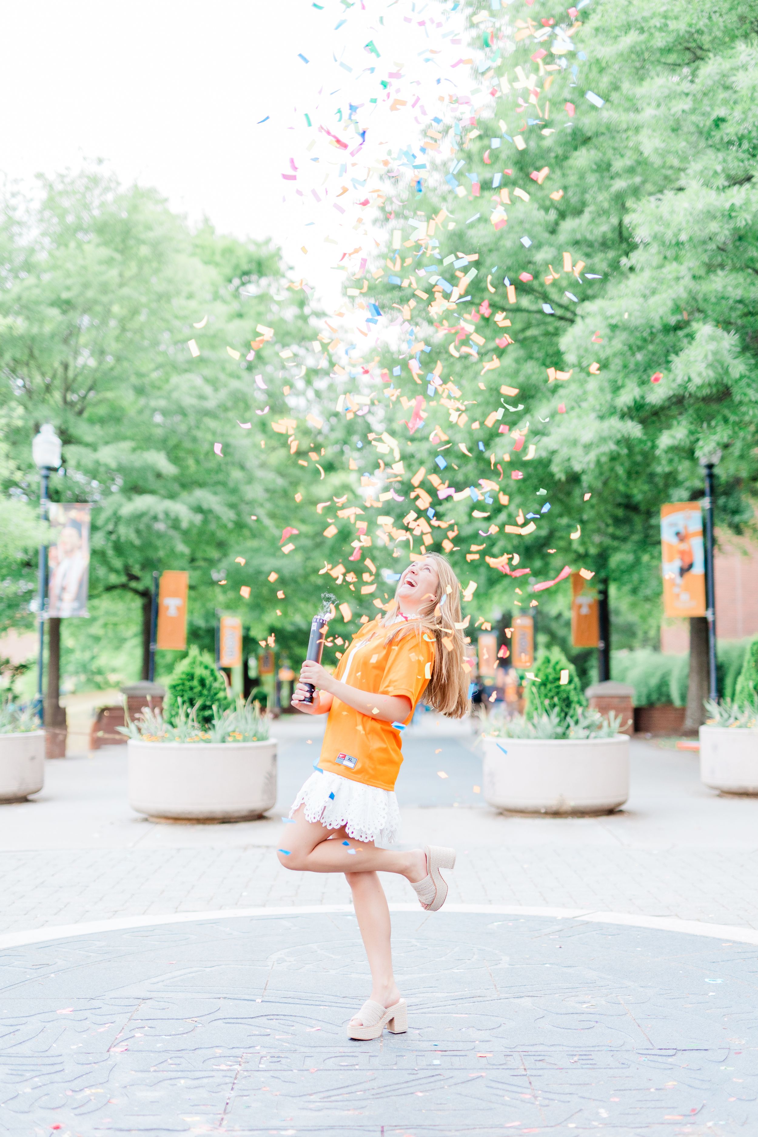 A person in orange clothing dances and twirls on a brick pathway surrounded by green trees and banners.