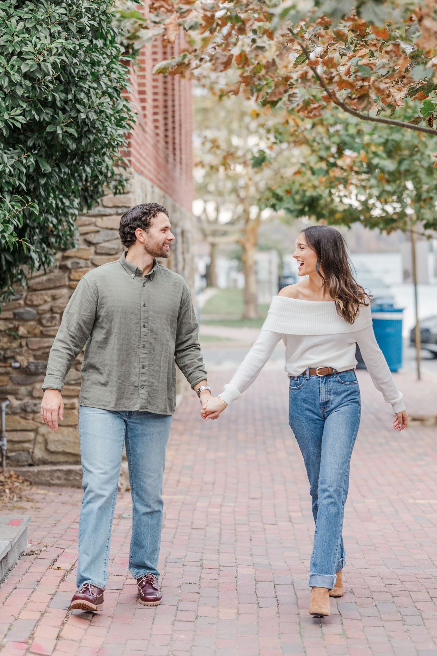 A couple in casual outfits holding hands and walking together on a brick pathway surrounded by autumn trees.