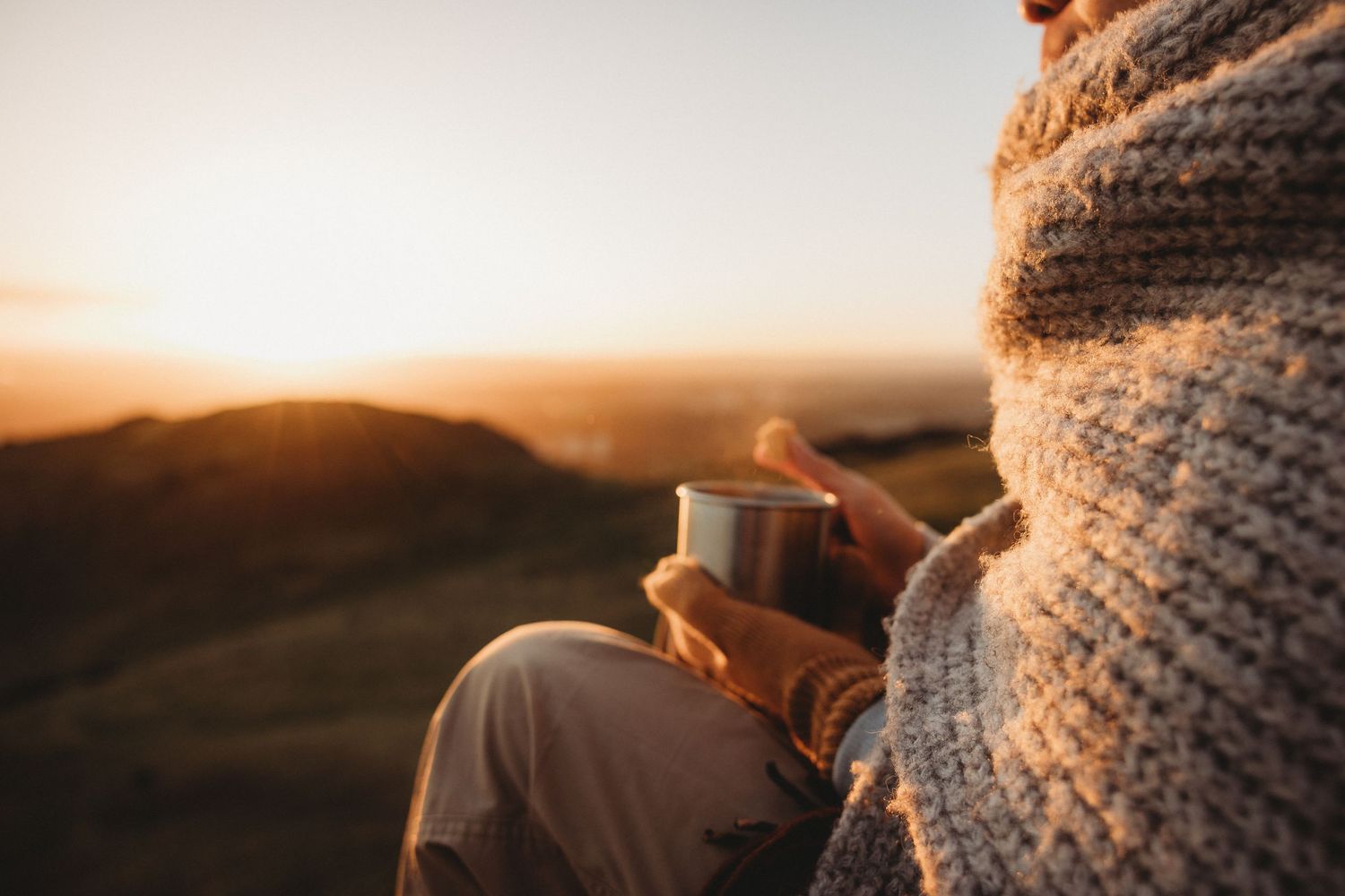 A cozy winter scene with hands holding a warm beverage in a knit sweater against a sunset backdrop.