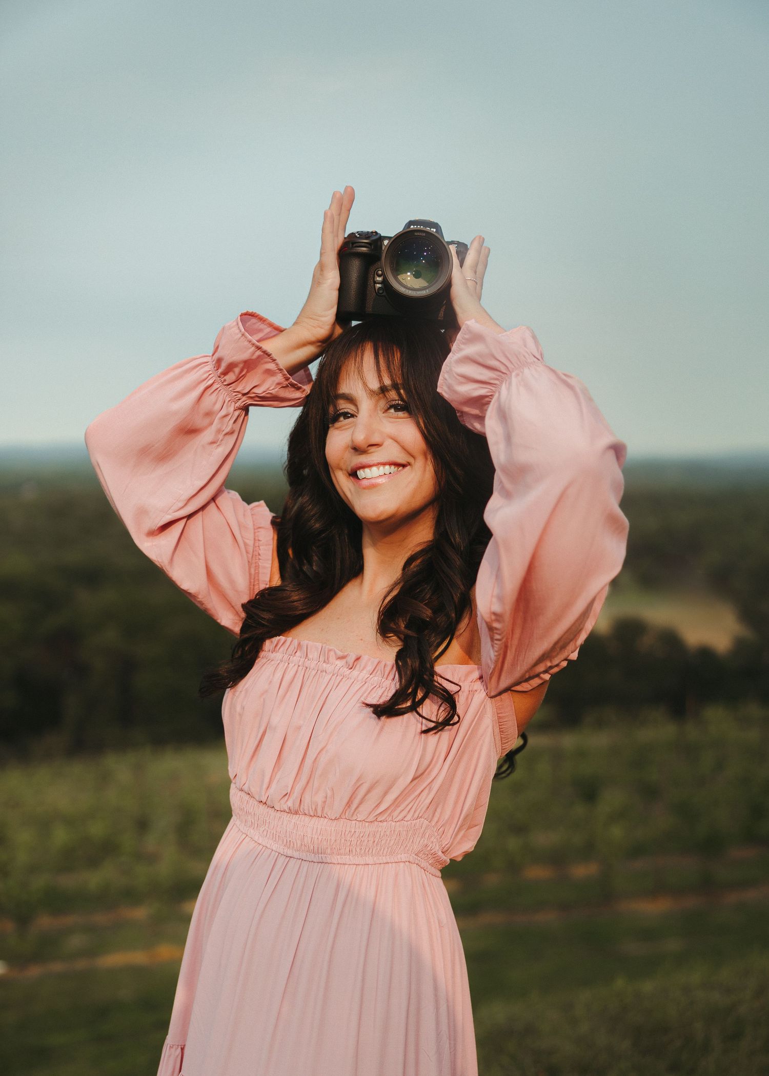 Woman in pink flowy dress poses playfully in outdoor field during sunset photoshoot.