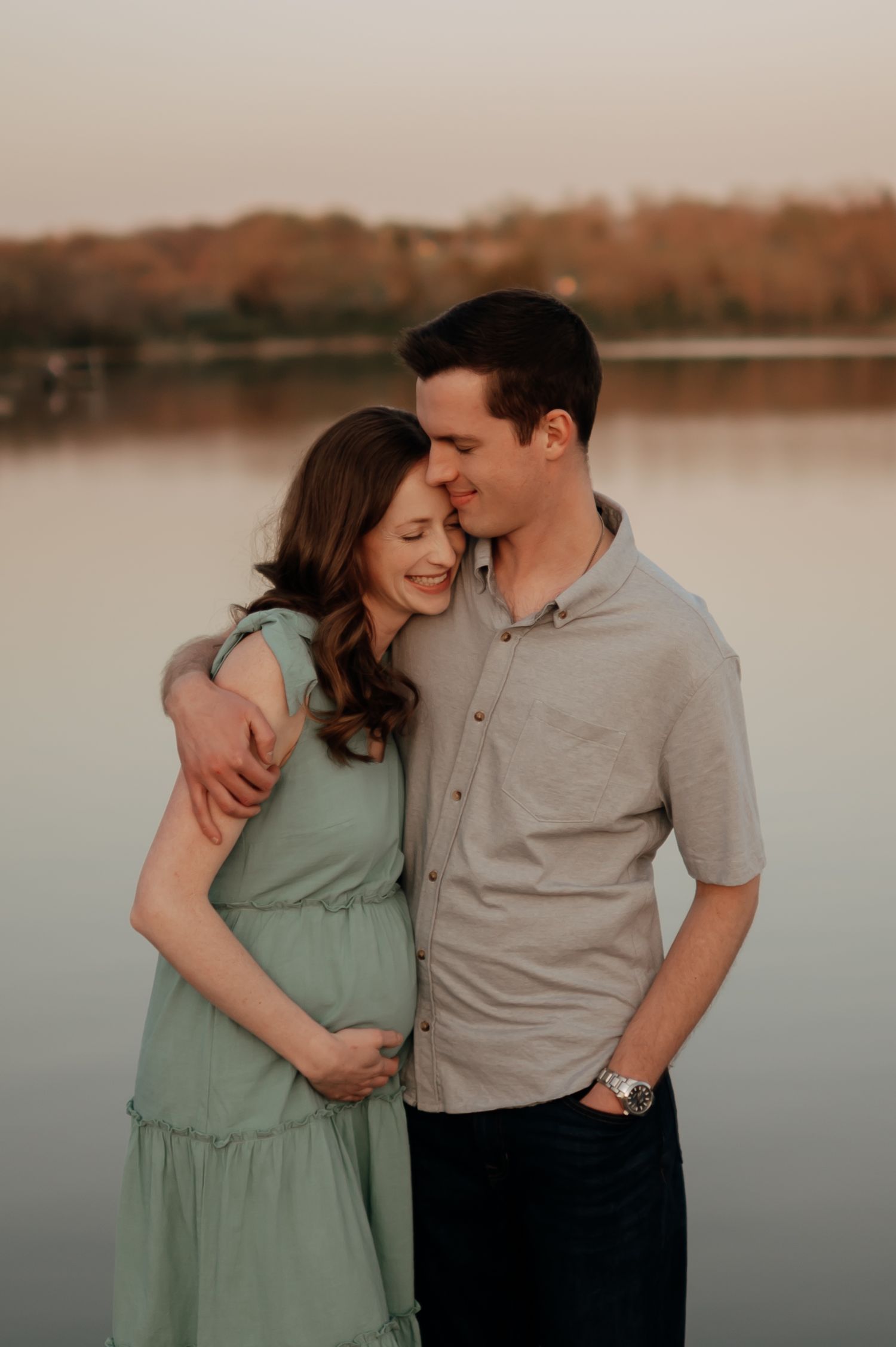 Couple sharing a tender moment by a lake at golden hour, woman in mint dress cradling baby bump.