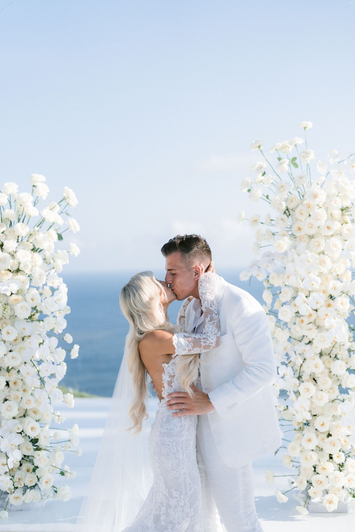 A romantic wedding ceremony by the sea with white floral arches and decorations against a blue ocean backdrop.