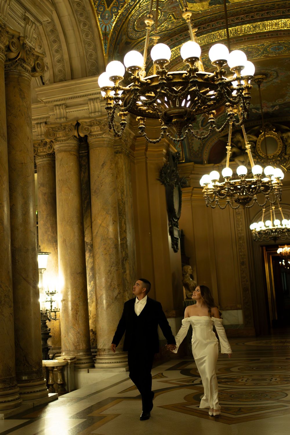 Elegant wedding couple walks through ornate hallway with marble columns and crystal chandeliers in historic building.