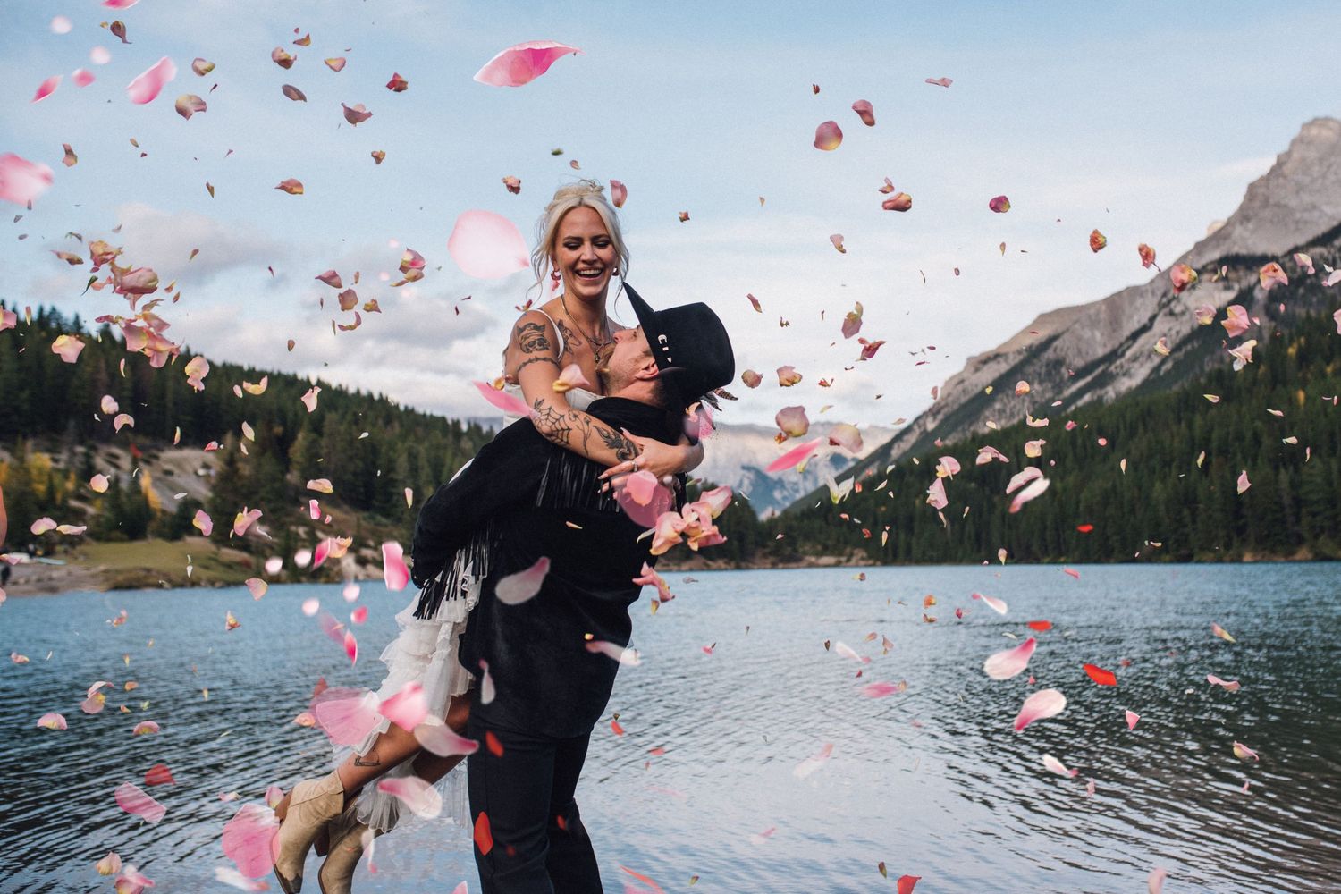 Pink flower petals float through the air as a couple embraces by a scenic mountain Banff lake on their wedding day.