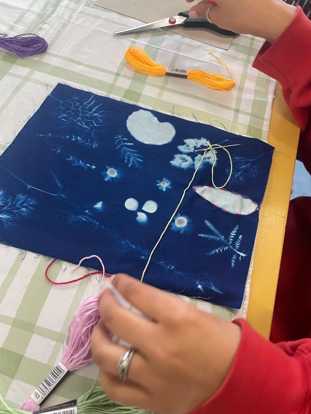 Someone in a red sleeve works on an embroidery project with a navy blue fabric featuring white floral cyanotype designs.
