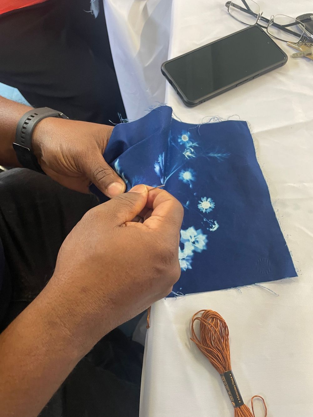 Hands working on blue floral fabric with embroidery thread and a smartphone visible on white surface.