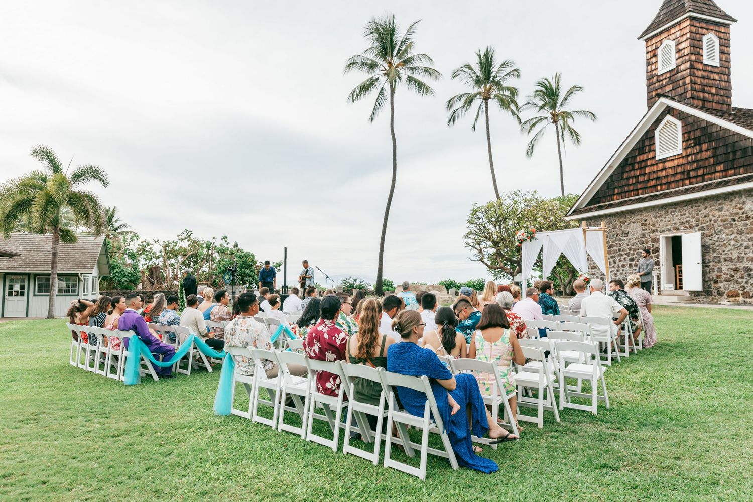 Outdoor wedding ceremony at a historic Hawaiian chapel with white chairs and palm trees against cloudy skies.