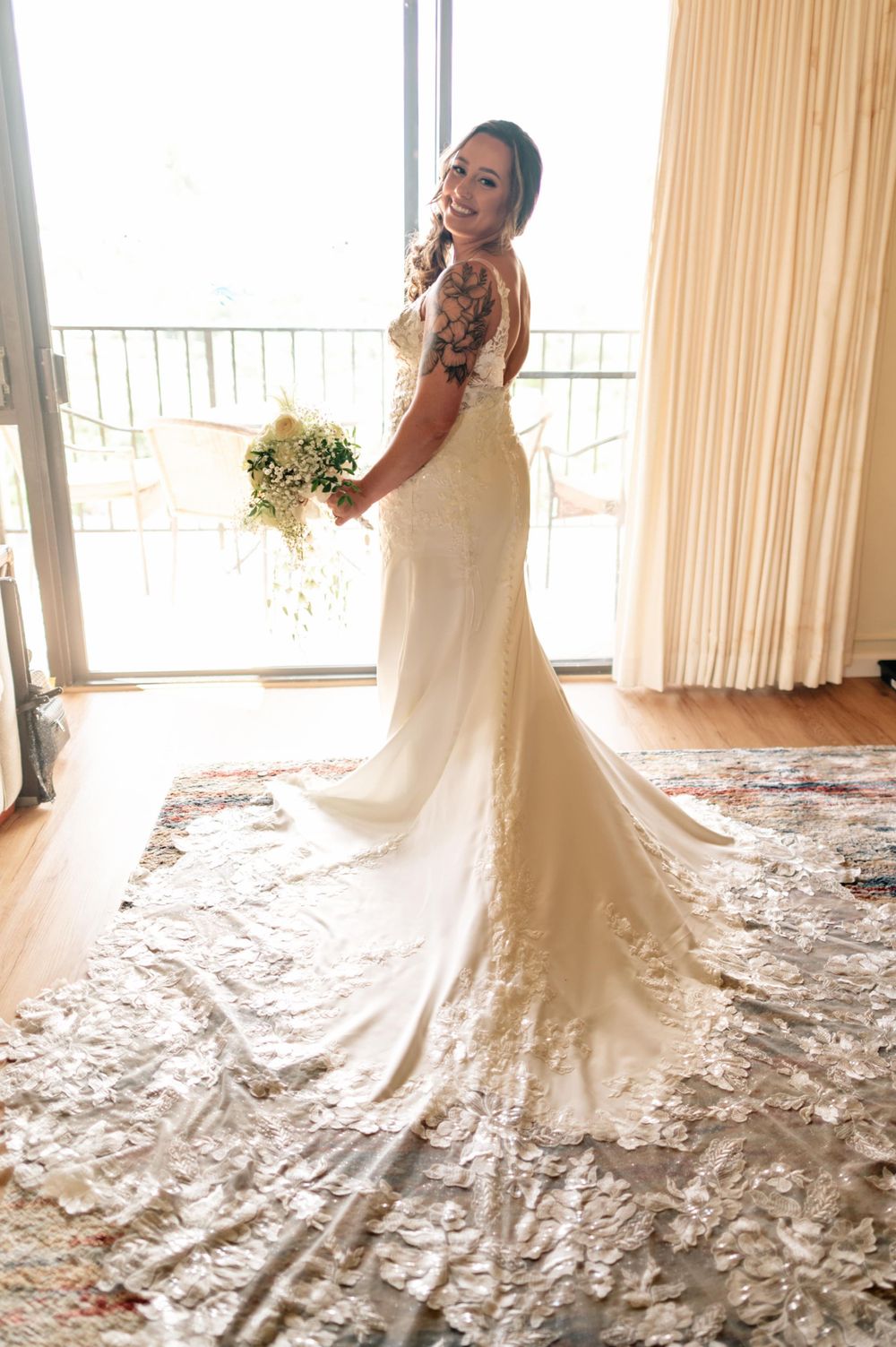 A bride in a fitted lace wedding gown with long train poses by a window with her bouquet.