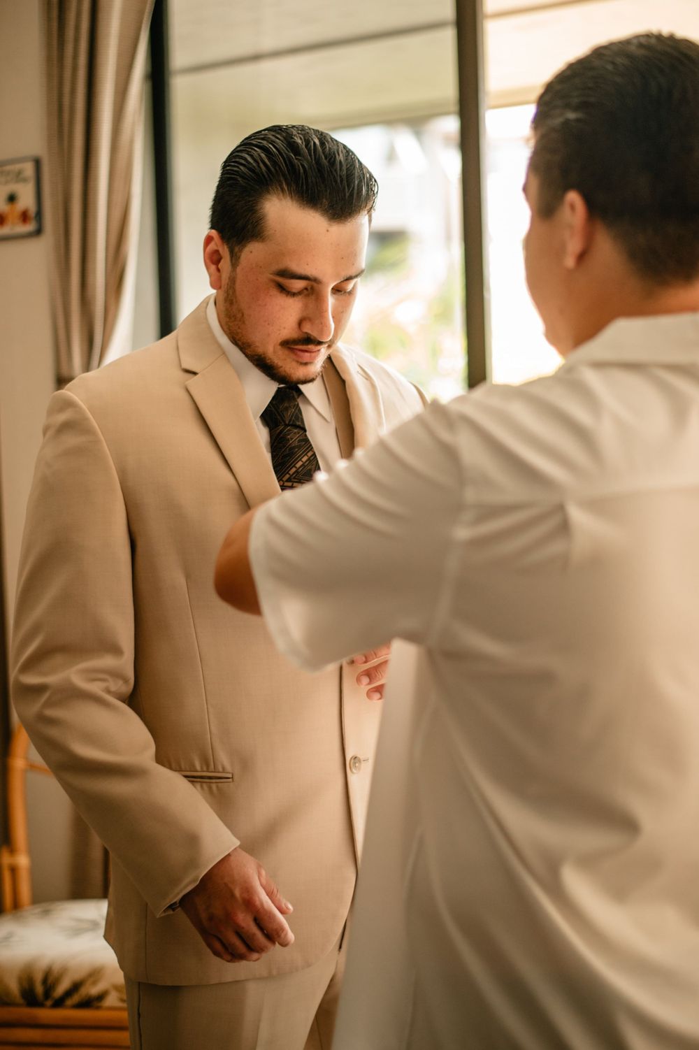 Groom getting dressed in a beige suit with help from others in a sunlit room.