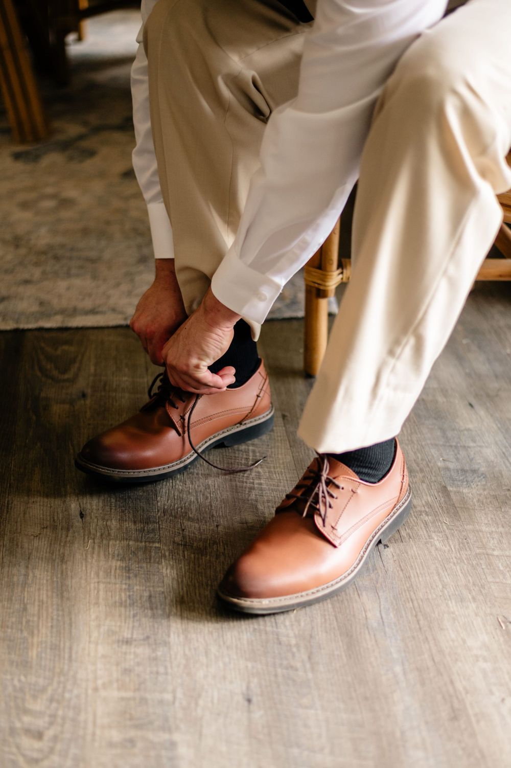 Someone lacing up brown leather dress shoes while wearing white formal attire.