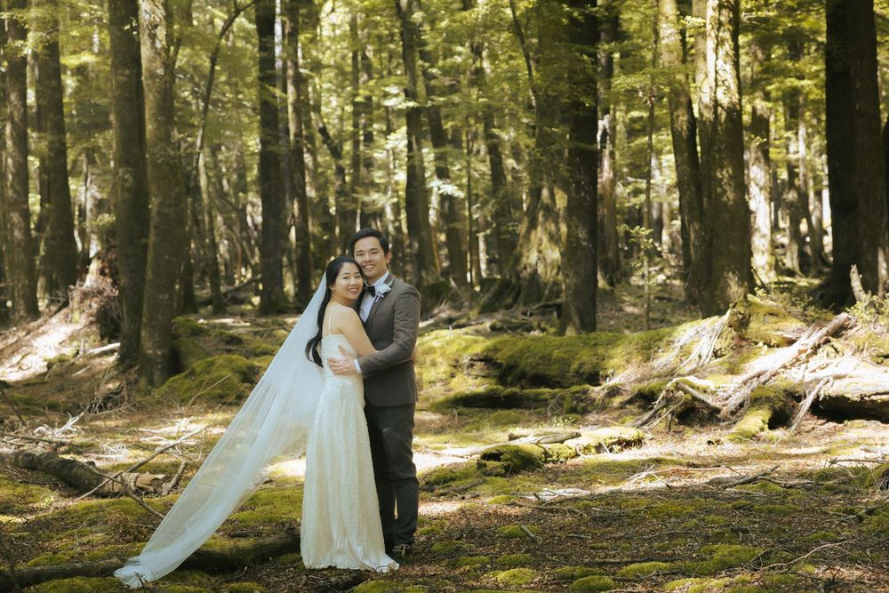 A couple in wedding attire stands in a sunlit forest clearing surrounded by tall evergreen trees.