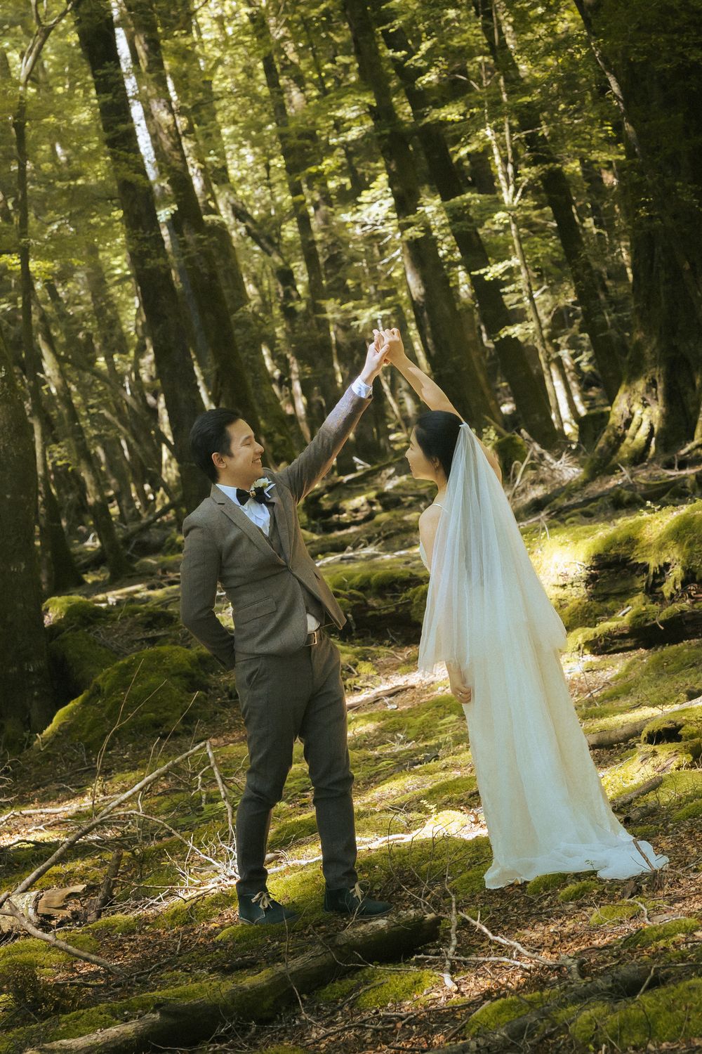 A couple dances playfully in a sunlit forest setting wearing wedding attire.