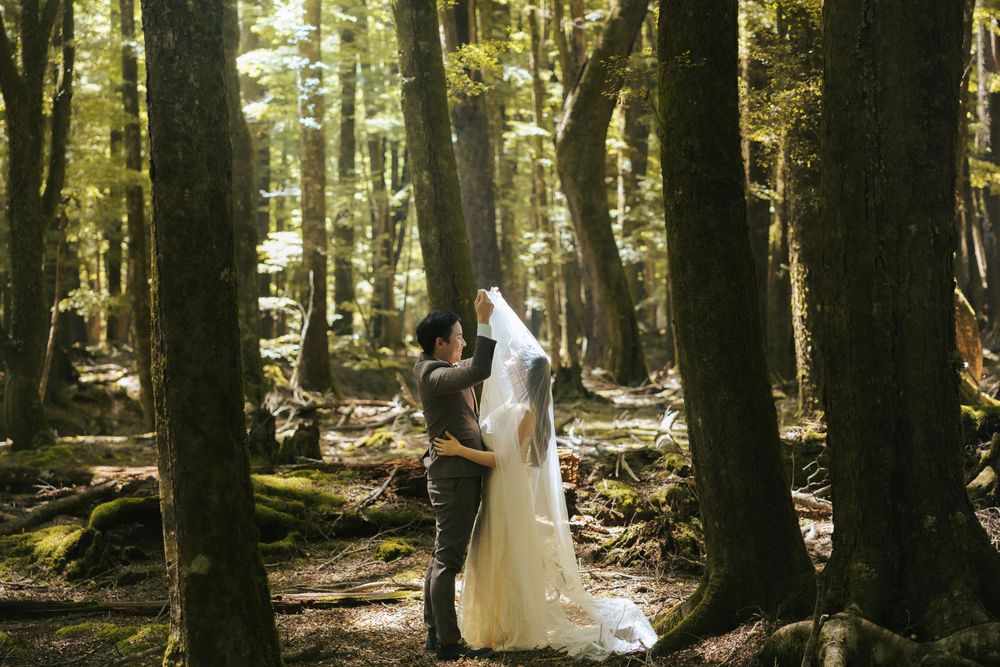 A couple in wedding attire poses in a sunlit forest with tall trees creating a magical woodland backdrop.