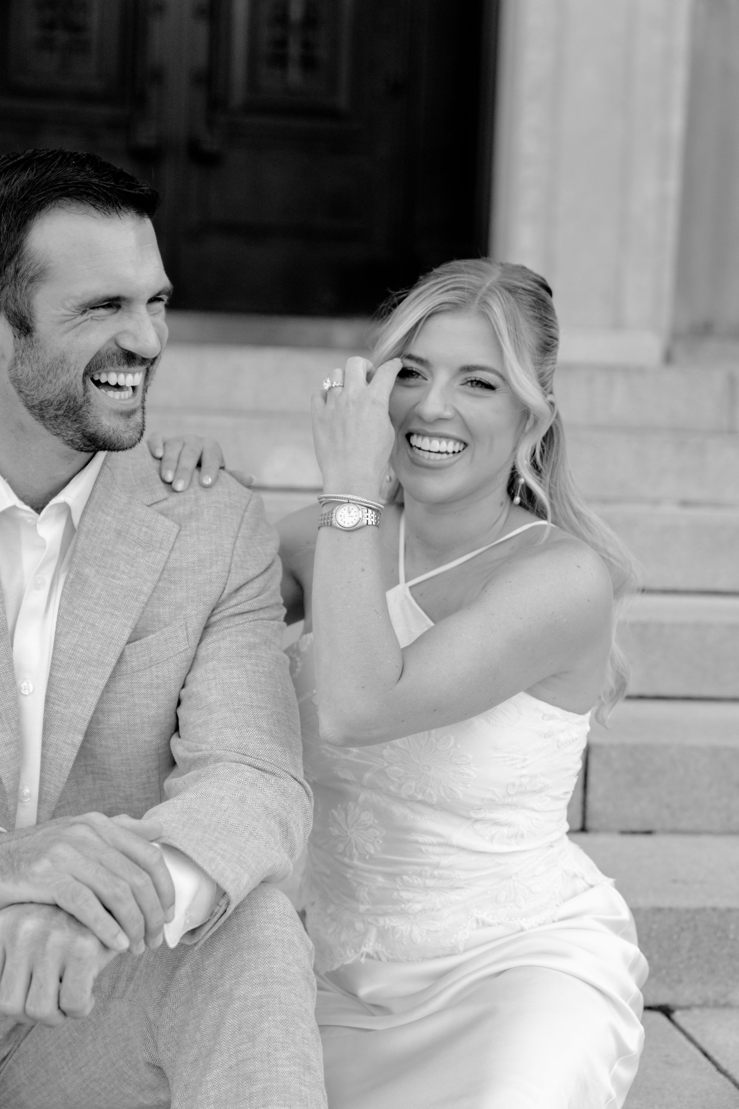 Black and white photo of a laughing bride and groom sitting on steps, wearing wedding attire.