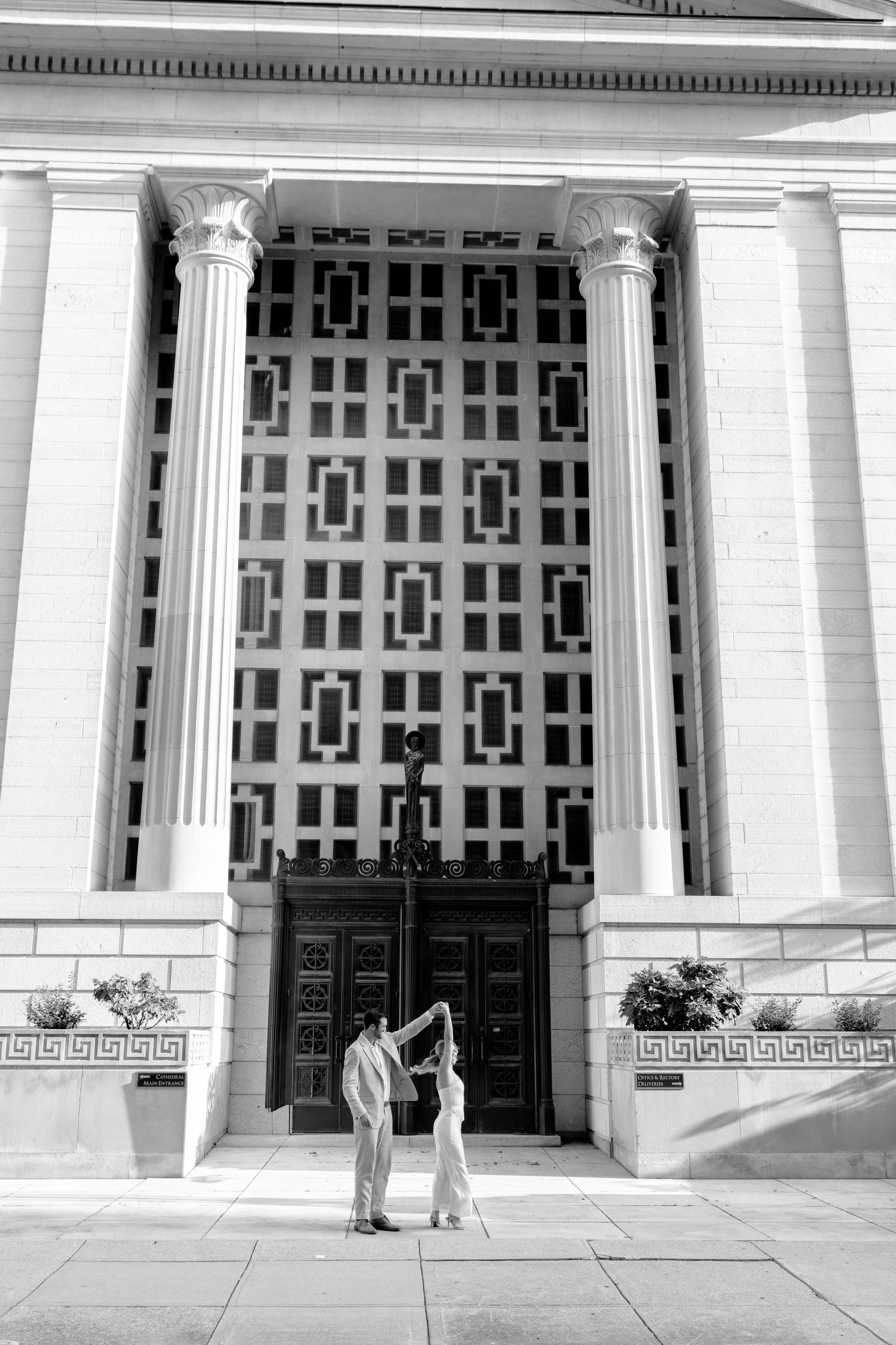 Black and white photo of a couple dancing in front of a grand neoclassical building with ornate doors.