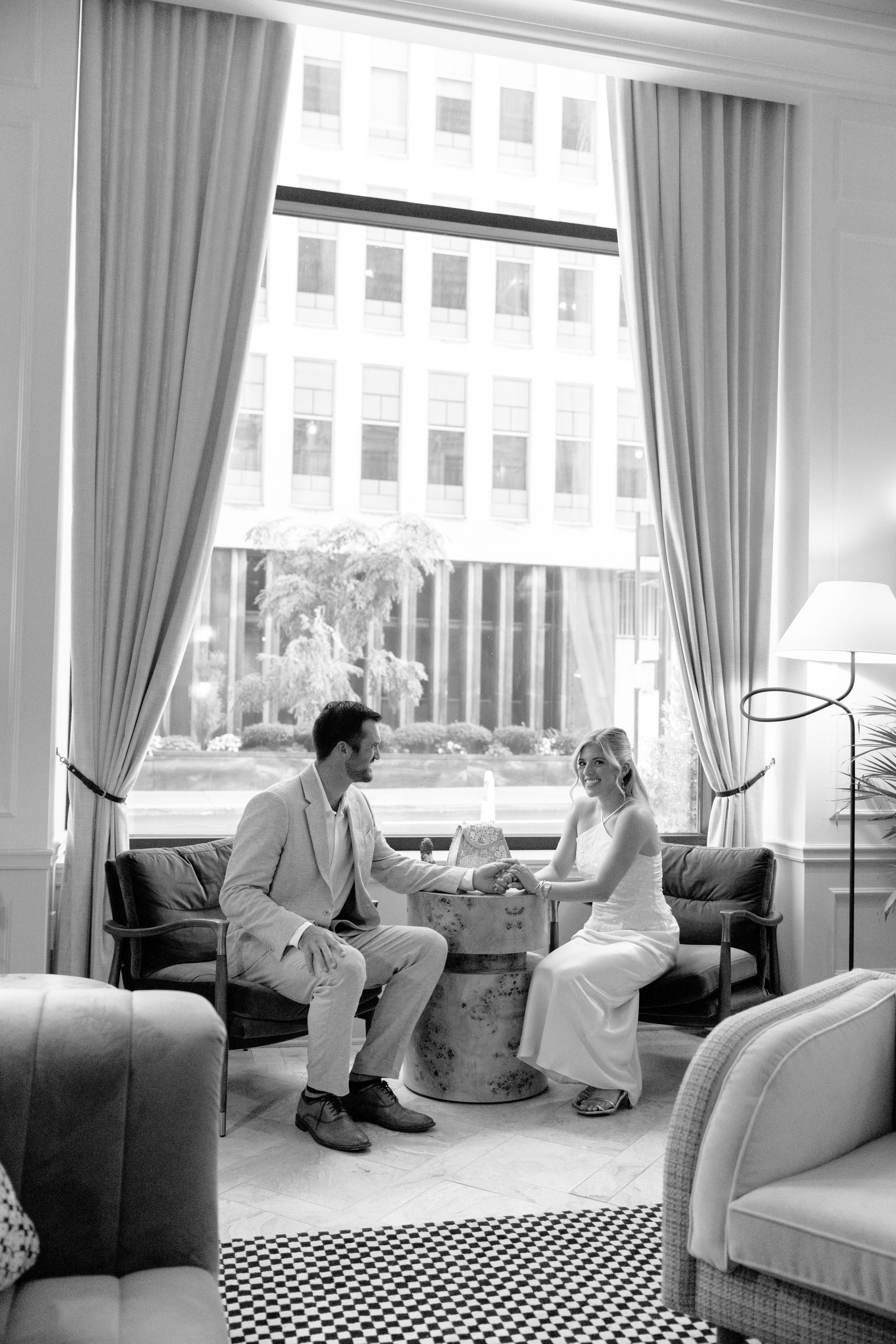 Black and white photo of a couple in wedding attire sitting at a small table by a large window in an elegant hotel lobby.