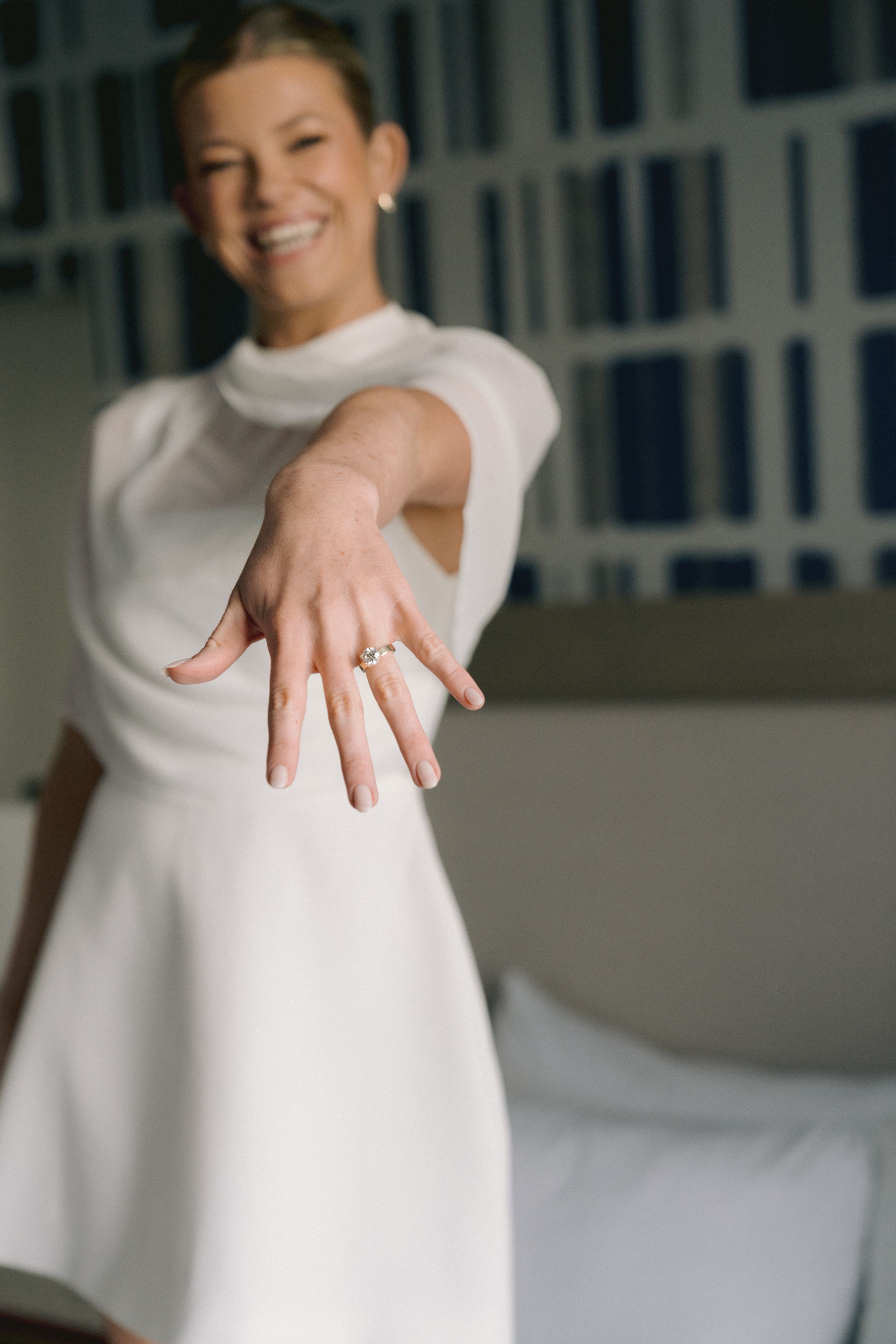 Smiling bride in white dress showing off her engagement ring by extending her hand toward the camera.